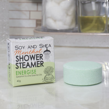 A boxed shower steamer and the steamer itself on a countertop in a bathroom setting. The box reads 'Soy and Shea Menthol Shower Steamer Energise' and the round steamer is a pale green color.
