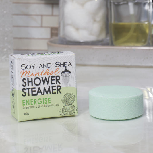 A boxed shower steamer and the steamer itself on a countertop in a bathroom setting. The box reads 'Soy and Shea Menthol Shower Steamer Energise' and the round steamer is a pale green color.