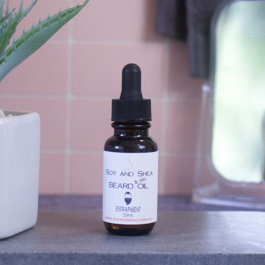 A brown bottle with white label and black dropper lid stands on stone surface next to a pot with green leaves against a blurred tiled background.  The label reads BEARD AND MO OIL and shows the image of a man with bead and moustache.