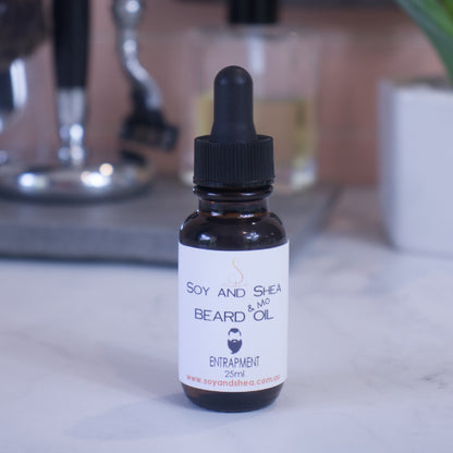A small brown bottle of "Soy and Shea Entrapment Beard & Mo Oil, 25ml" sits on the countertop. In the background, are blurred  shaving accessories and a small plant against a peach background.
