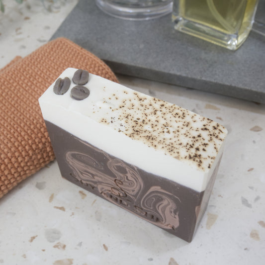 A bar of soap sits on a marble counter next to a orange cloth.  The soap is dark brown with tan swirls and has a white layer on top.  The top is decorated with brown speckles and 3 coffee beans made of soap