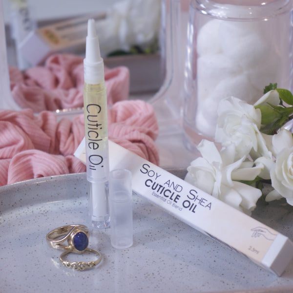 A clear, narrow tube with the lid removed showing a brush tip stands upright on a stone tray.  Beside it sits a cardboard box labelled CUTICLE OIL and a set of gold rings.  In the background is a bathroom counter top with a pink scrunchie, small mirror, jar of cotton balls and a some white  flowers.