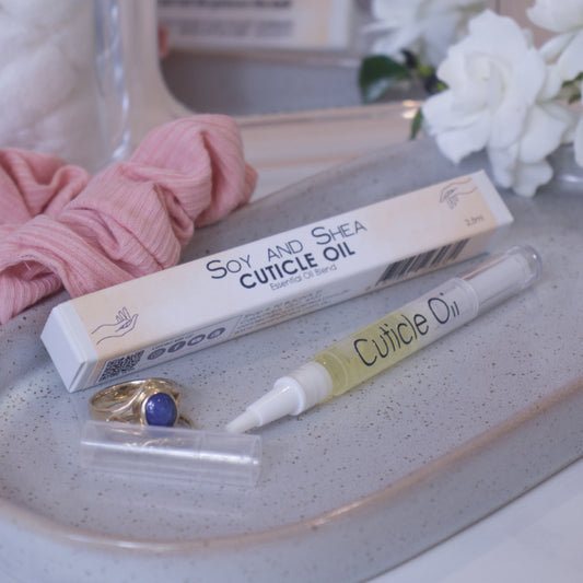A clear, narrow tube with the lid removed showing a brush tip lies on a stone tray beside a cardboard box labelled CUTICLE OIL and a set of gold rings.  In the background is a bathroom counter top with a pink scrunchie, small mirror, jar of cotton balls and a some white  flowers.