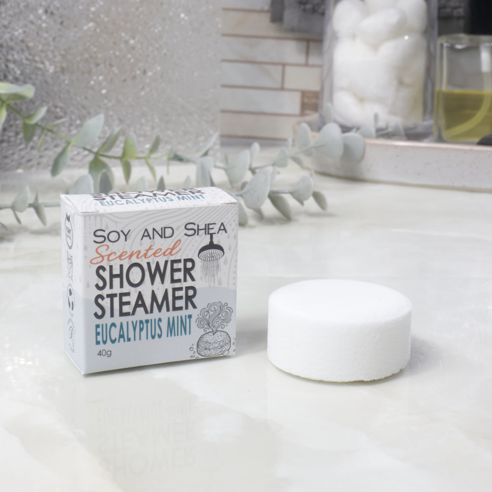 A boxed scented shower steamer and the steamer itself on a bathroom counter. The box reads 'Soy and Shea Scented Shower Steamer Eucalyptus Mint', and the round white steamer sits beside it.
