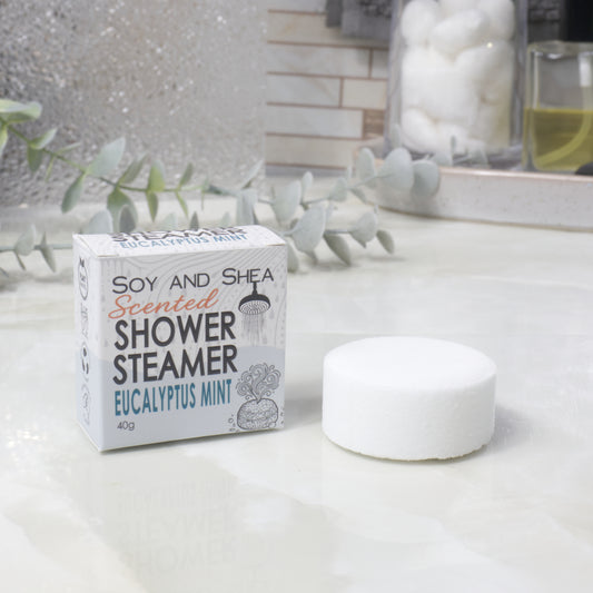 A boxed scented shower steamer and the steamer itself on a bathroom counter. The box reads 'Soy and Shea Scented Shower Steamer Eucalyptus Mint', and the round white steamer sits beside it.