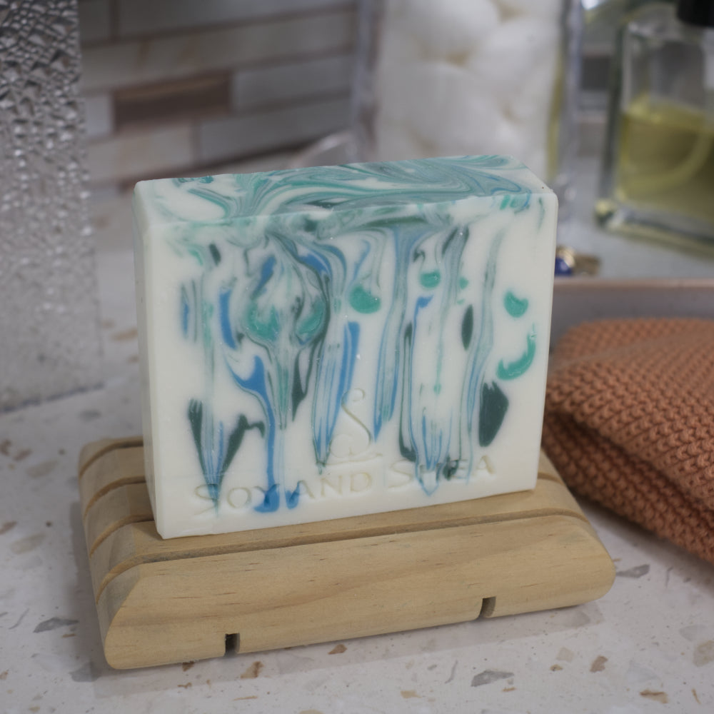 A white bar of soap with blue and green drops of colour sits on a wooden dish on a bathroom counter