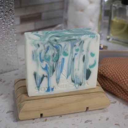 A white bar of soap with blue and green drops of colour sits on a wooden dish on a bathroom counter