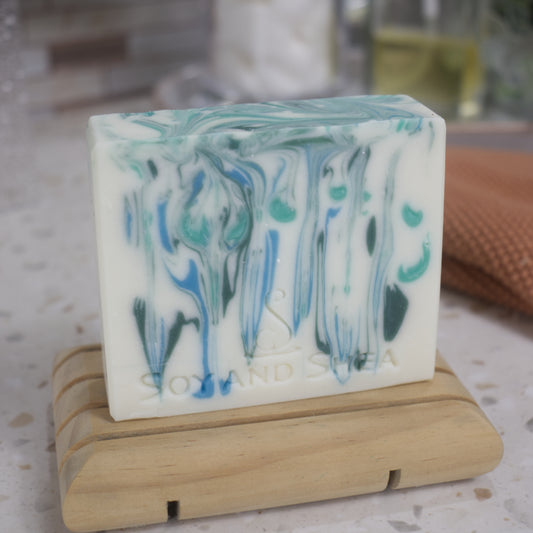 Marbled soap bar on a wooden stand with a blurred background.  The soap has a white background with drips of blue and green throughout