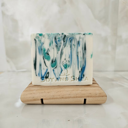 A white bar of soap with blue and green drops of colour sits on a wooden stand surrounded by marble tiles