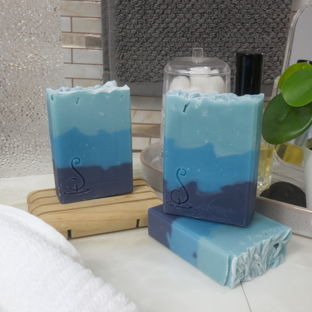Three bars of soap are presented on a bathroom counter top next to a white towel and some accessories.  One soap lays flat on the marble top while another stands upright on on it.  Behind and to the left is a third upright bar on a wooden soap rack.  All soaps feature three layers of blue colour starting with the darkest the bottom and lightest on top
