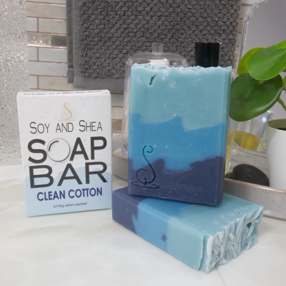 A selection of soap bars and their packaging is displayed on a bathroom countertop.  The front right, a soap bar lays flat on the marble top while a second bar stands upright on top  of it.  Behind and to the left is the box labeled as Soap Bar Clean Cotton.  The soaps have 3 layers of colour ranging from dark blue on the base and light on the top.
