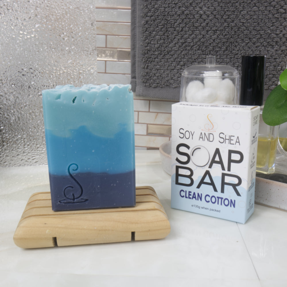 On a bathroom countertop, a tall rectangular soap stands on a wooden soap rack next to its packaging identifying it as Clean Cotton.  The soap has three layers of colour  starting with dark blue on the base, a medium blue through the middle and  a light blue on top.  