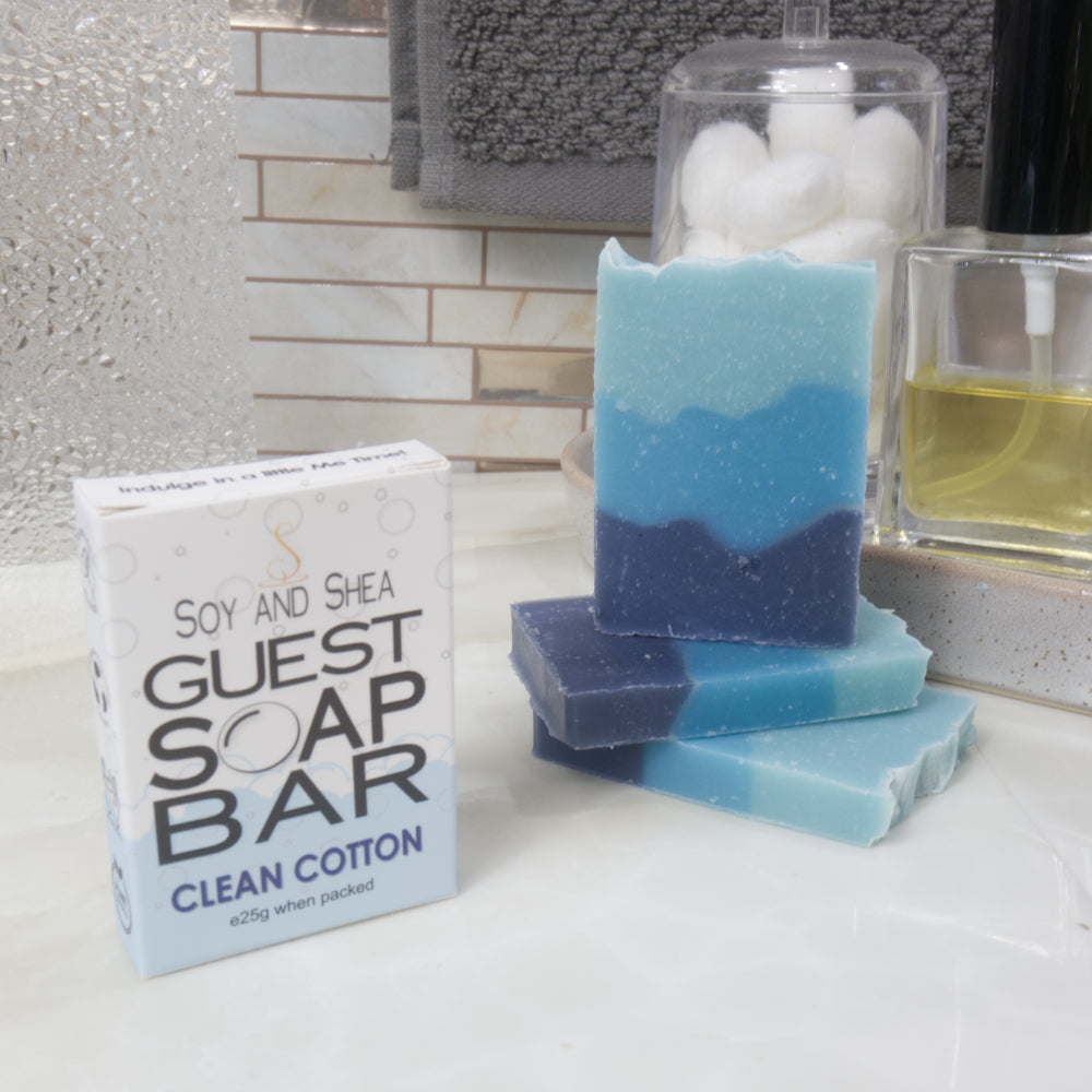A boxed bar of soap labeled 'Guest Soap Bar Clean Cotton' sits beside three stacked handmade soap bars with a blue and navy color gradient, set on a bathroom counter with towels and toiletries in the background.
