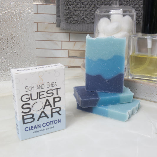 A boxed bar of soap labeled 'Guest Soap Bar Clean Cotton' sits beside three stacked handmade soap bars with a blue and navy color gradient, set on a bathroom counter with towels and toiletries in the background.