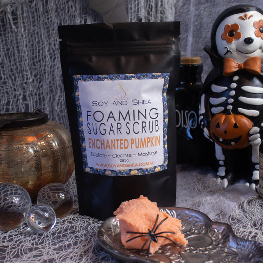 A Halloween-themed scene showcases Soy and Shea's Enchanted Pumpkin Foaming Sugar Scrub (Halloween Edition), enriched with Kaolin clay, elegantly packaged in a black pouch. The scrub is surrounded by a pumpkin, a skeleton figure, glass spheres, and cobwebs, creating a spooky and festive atmosphere.