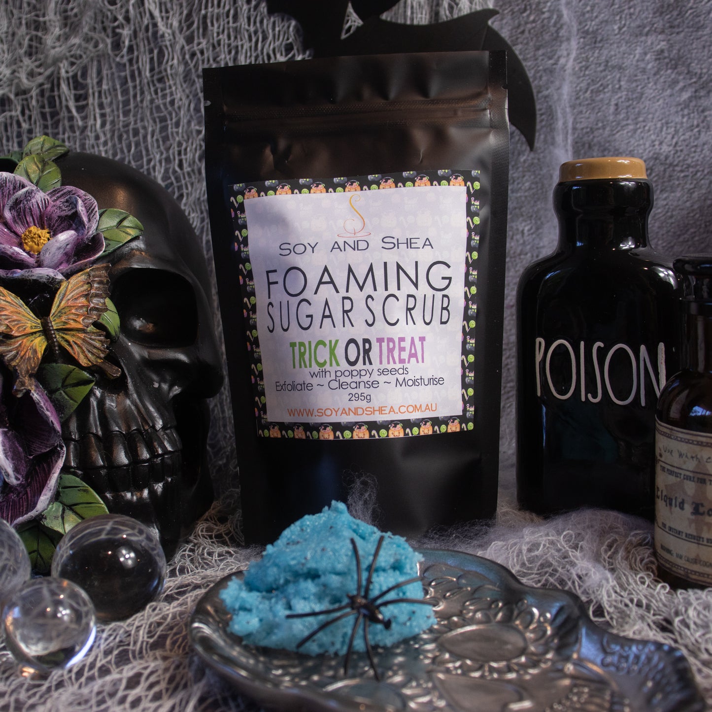 A Halloween-themed scene features a bag of exfoliating Trick or Treat Foaming Sugar Scrub (Halloween Edition) from Soy and Shea on a cobweb-covered surface. The bright blue scrub, adorned with a spider decoration, is surrounded by a black skull, a "Poison" bottle, a potion bottle, and small glass orbs. This spooky setup emphasizes the scrub's eerie allure.