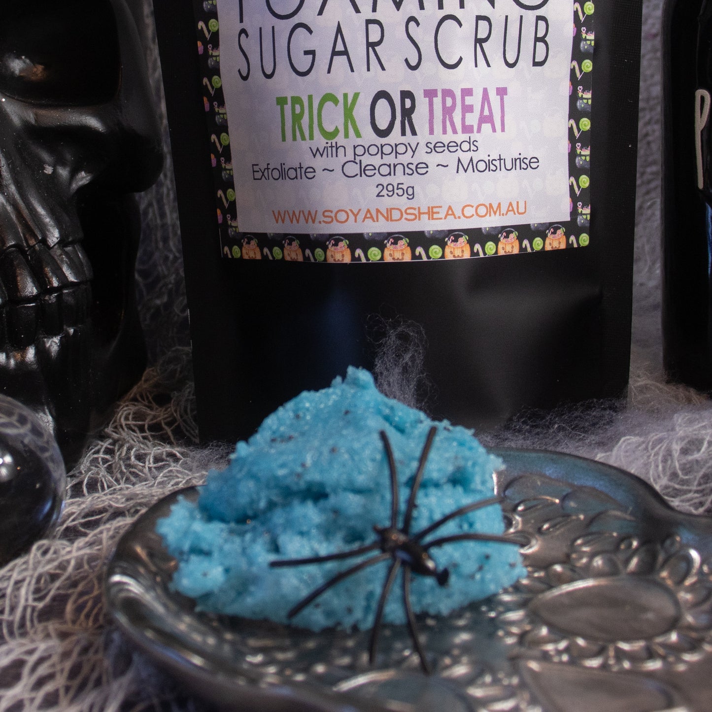 A jar labeled "Trick or Treat Foaming Sugar Scrub (Halloween Edition)" from Soy and Shea is surrounded by Halloween-themed decor. In the foreground, the blue exfoliating scrub featuring a black spider figurine sits on a silver dish, enriched with nourishing cottonseed oil for a spooky yet pampering experience.