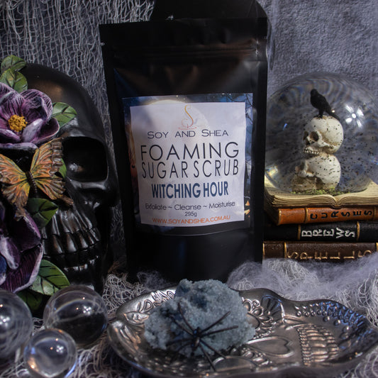 A black pouch labeled "Witching Hour Foaming Sugar Scrub (Halloween Edition)" from Soy and Shea is surrounded by Halloween-themed items, including a black skull, dried flowers, a silver tray with a blue crystal, and old books. This exfoliating scrub is enriched with cottonseed oil to offer an enchanting skincare experience.