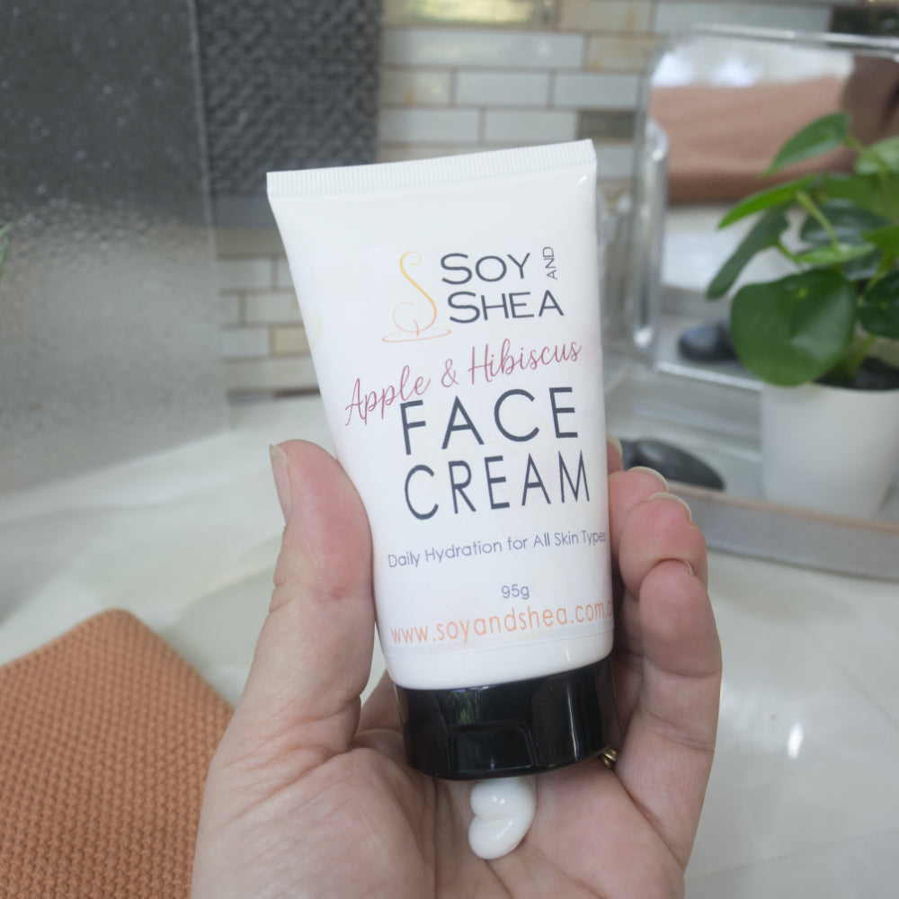 A hand holding a white squeeze tube of Soy and Shea Face Cream with Apple & Hibiscus flavor, set against a softly blurred bathroom background with a plant and mirror.  A small amount of white cream sits in the palm of the hand