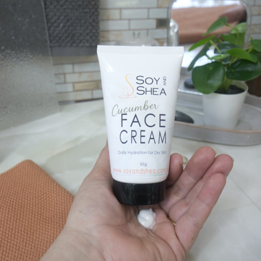 A hand holding a white squeeze tube of Soy and Shea Cucumber Face Cream, with a small dollop of white cream resting on the palm. The background shows a softly blurred bathroom setting with a sink, tiled wall, and a potted plant.