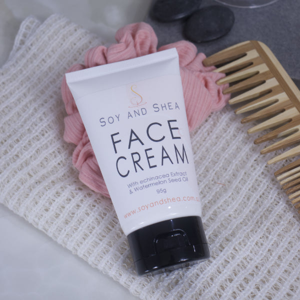 A white tube with black lid of Soy and Shea Face Cream rests against a pink scrunchie on top of a cotton cloth.  Next to it is a wooden comb