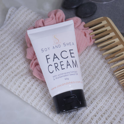 A white tube with black lid of Soy and Shea Face Cream rests against a pink scrunchie on top of a cotton cloth.  Next to it is a wooden comb