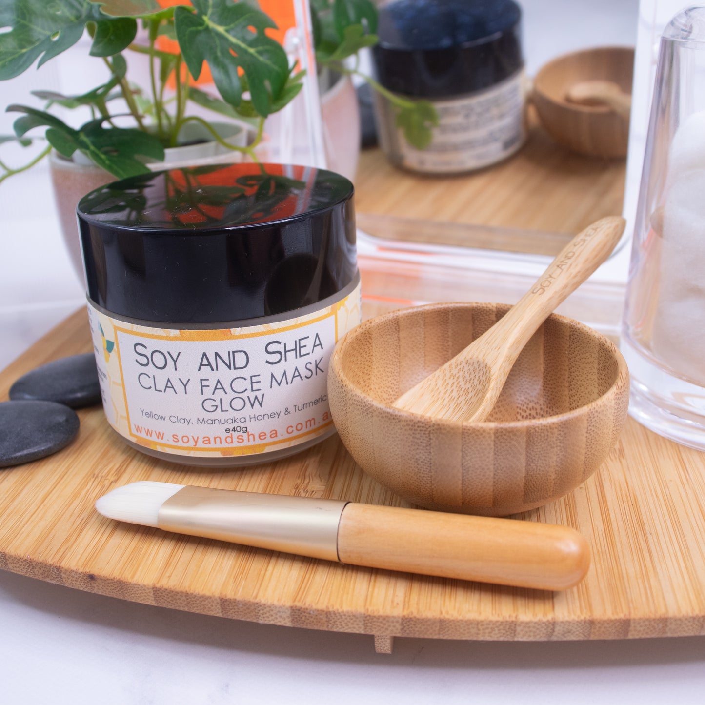 A jar of the Soy and Shea Clay Face Mask Glow sits on a wooden tray alongside items from the Clay Face Mask Utensil Set, which includes a small mixing bowl, a bamboo spoon, and a face mask brush. In the background, leaves and black stones are visible along with cotton pads in a clear container.