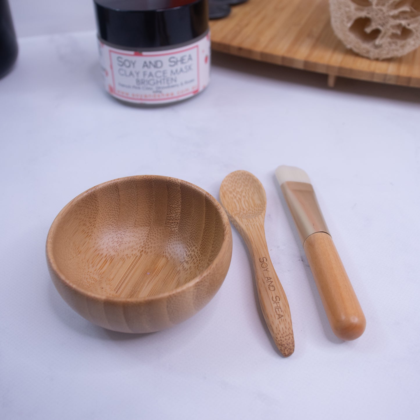 A wooden bowl, a bamboo spoon, and a face mask applicator brush from the Clay Face Mask Utensil Set are arranged on a white surface. Behind them is a jar labeled "Soy and Shea Face Mask" in red and white. A wooden board and a natural sponge are partially visible in the background.