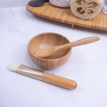 A small wooden bowl from the Soy and Shea Clay Face Mask Utensil Set, complete with a matching bamboo spoon, is placed on a white surface. Nearby lies a makeup brush featuring light-colored bristles and a wooden handle. In the background, a wooden tray holds a natural sponge, skincare product, and what appears to be a clay face mask from the Soy and Shea collection.