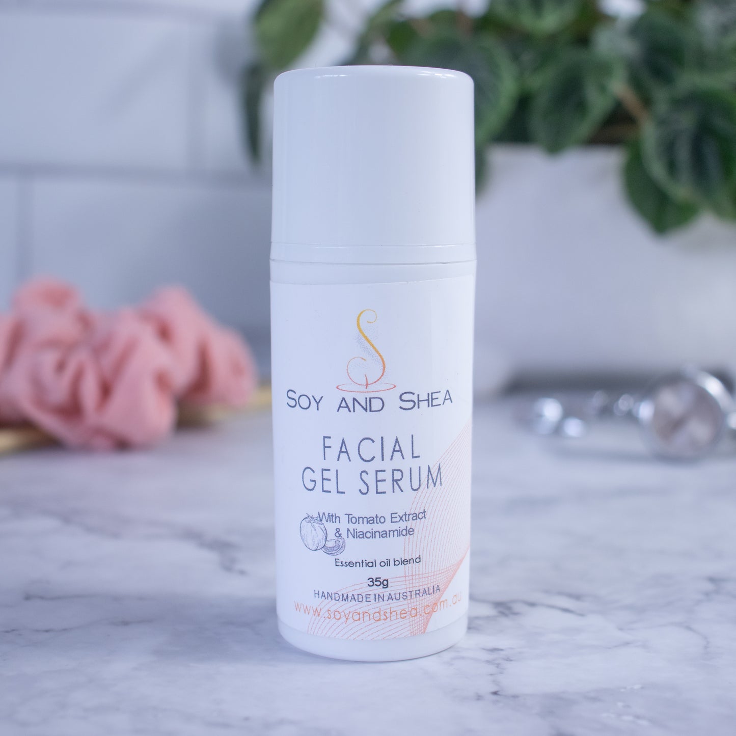 A white bottle of the Soy and Shea Facial Gel Serum sits on a marble surface with a blurred background of a plant, scrunchie and watch.  The bottle has a white cap and the label has red highlights