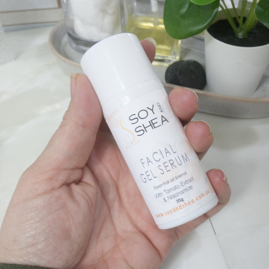 A hand holding a white cylindrical bottle of Soy & Shea Facial Gel Serum, with a marble countertop and potted plants in the blurred background.