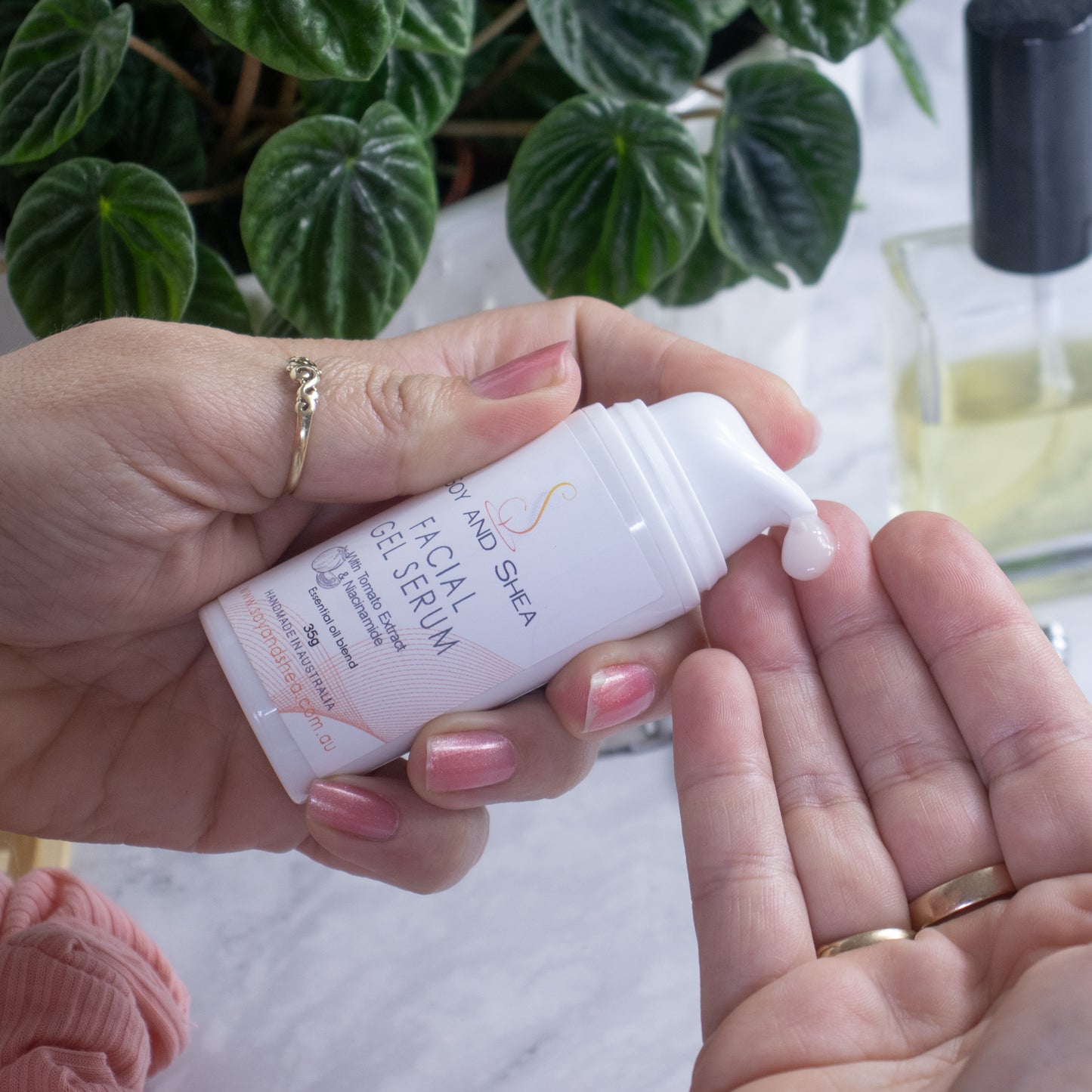 A hand holding a white bottle with serum pump labeled "Soy and Shea Facial Gel Serum" dispenses a small amount of white liquid onto the fingertips of another hand. In the bright background, there's a plant with green leaves and a perfume bottle. 