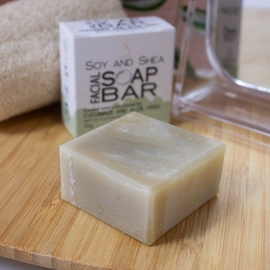 A close up image of a smalll squarish bar of soap in a pale green colour lays flat on a wooden surface. Behind is a white box with green bubble foam border reading FACIAL SOAP BAR next to a mirror and loofah