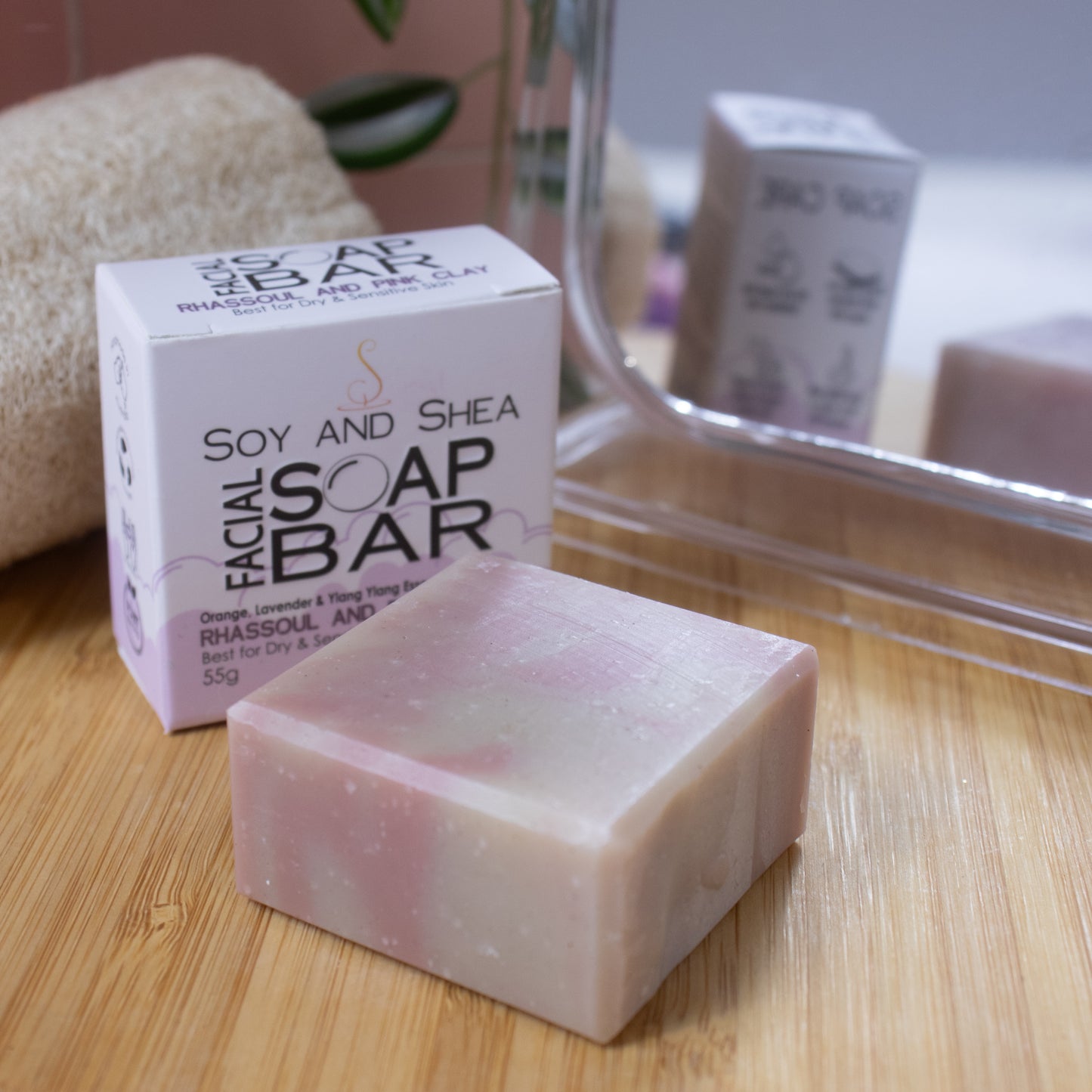 A close up image of a smalll squarish bar of soap in a pale pink and cream colours lays flat on a wooden surface. Behind is a white box with green bubble foam border reading FACIAL SOAP BAR next to a mirror and loofah