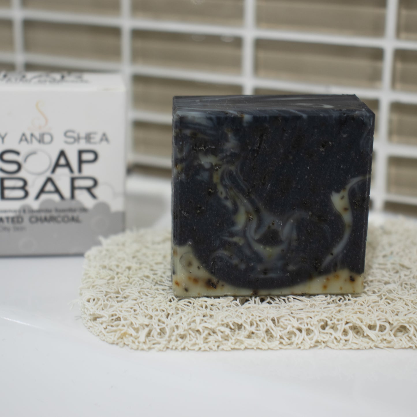 A small square bar of soap sits on a cream soap mat on white surface agains a brown tile with a SOAP BAR packaging box in the background.  THe soap is black with wisps of pale green and brown flecks