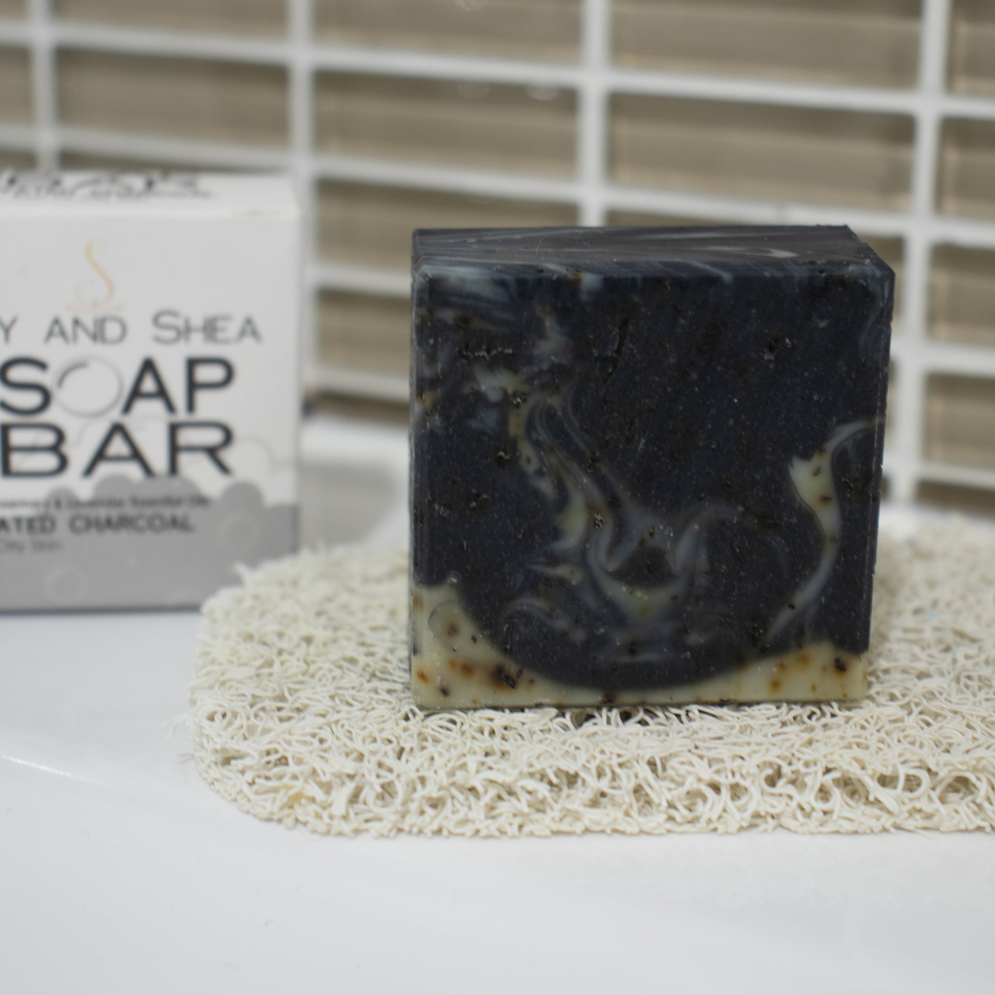 A small square bar of soap sits on a cream soap mat on white surface agains a brown tile with a SOAP BAR packaging box in the background.  THe soap is black with wisps of pale green and brown flecks
