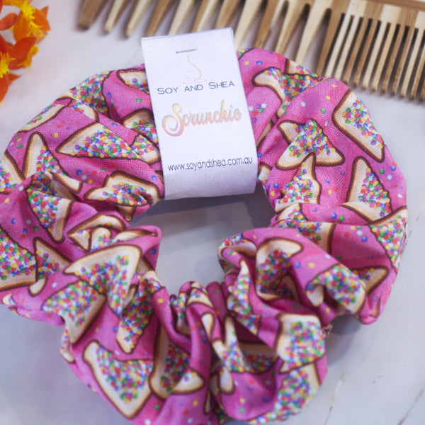 A pink, handmade cotton scrunchie with a toast and colorful sprinkles pattern features a knotless elastic design. The tag reads "Soy and Shea Fairy Bread Scrunchie" along with the website address, set against an elegant backdrop of wooden combs and vibrant orange flowers.