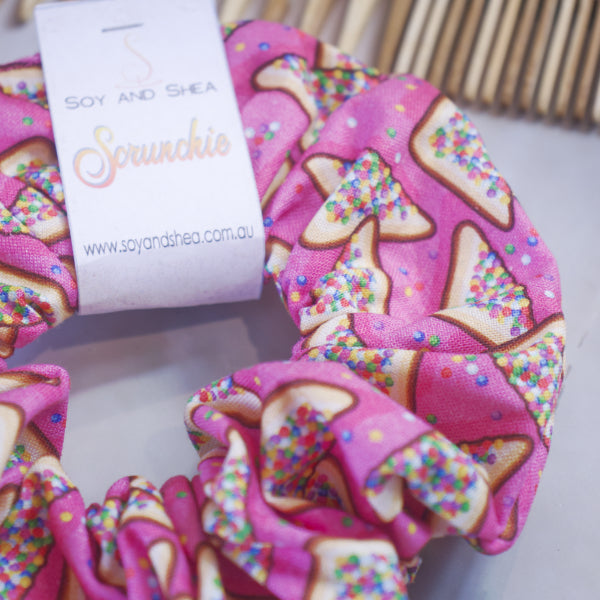 The "Fairy Bread Scrunchie" by Soy and Shea is a vibrant pink accessory adorned with a toast and sprinkles pattern, featuring a knotless elastic design for added style and comfort.