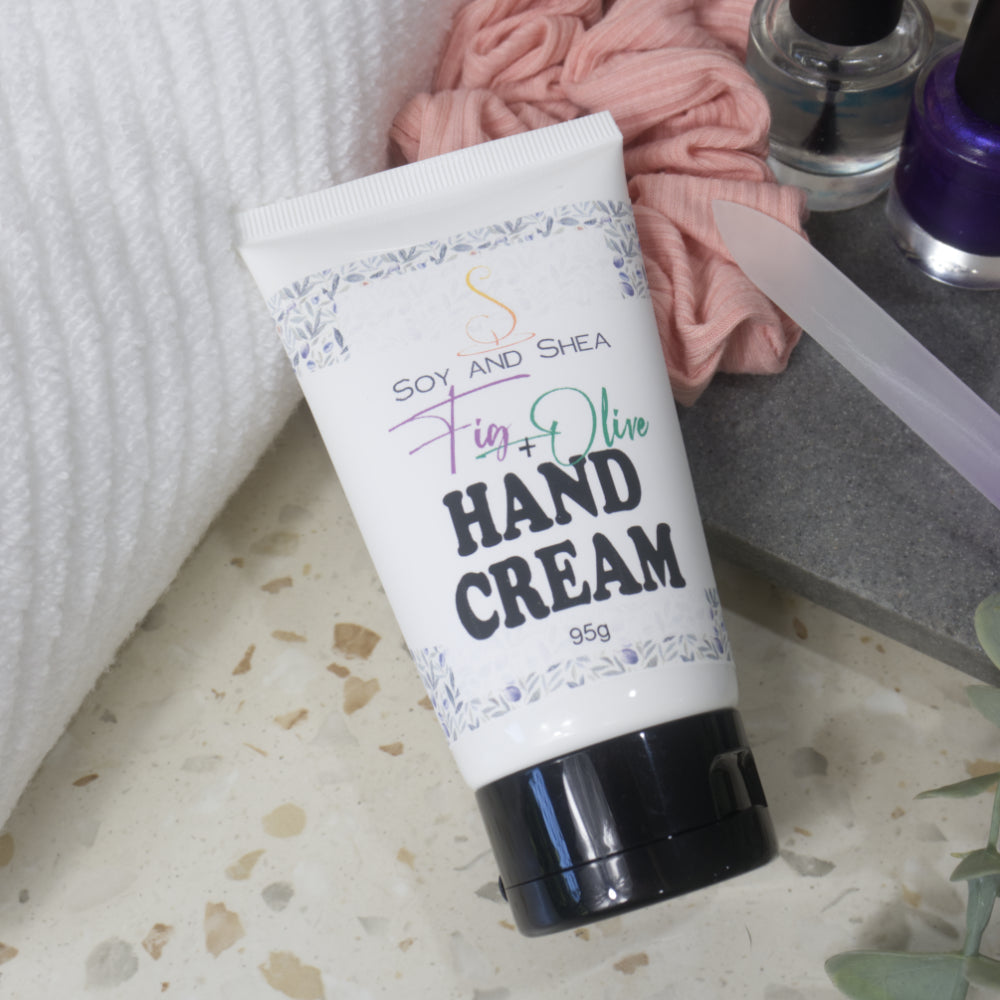 A white tube with black flip top lid lyes across a terrazzo surface, propped up against a white towel and a scrunchie that rests on a grey tray.  Two bottles of nail polish can be seen in the background along with nail file.  The tube features a product label identifying it has Fig & Olive Hand Cream and features a decorative border on the label of olive leaves and figs