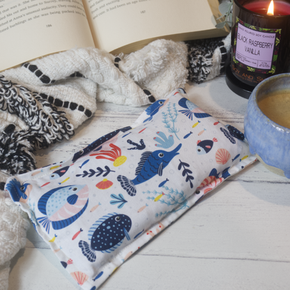 A small rectanglular Heat Pack rests on a wooden table top along with a black and white blanket, open book, burning candle and filled coffee cup evoking a cosy scene.  The Heat pack has a white background covered with illustrations of a variety of reef fish and coral in shades of blue.  