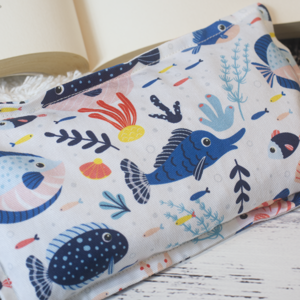 A close up image of the Mini Fish Heat pack highlighting the fabric design which shows illustrations of a variety of reef fish in shades of blue  along with pieces of coral on a white background.