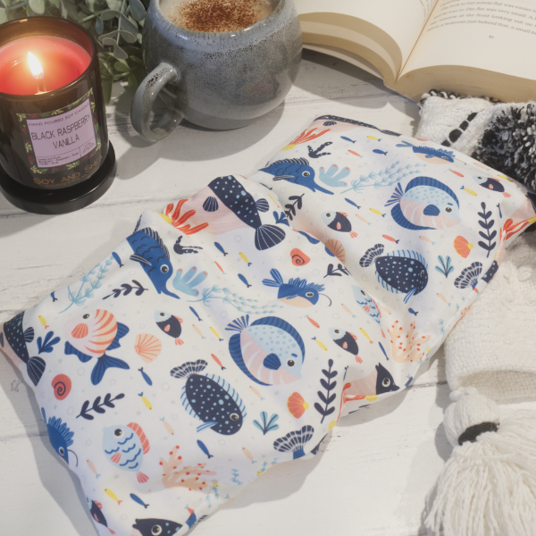 A small rectanglular Heat Pack rests on a wooden table top along with a black and white blanket, open book, burning candle and filled coffee cup evoking a cosy scene.  The Heat pack has a white fabric with a variery of reef fish in shades of blue with hinds of orage.  The fish are surrounded by pieces of coral.