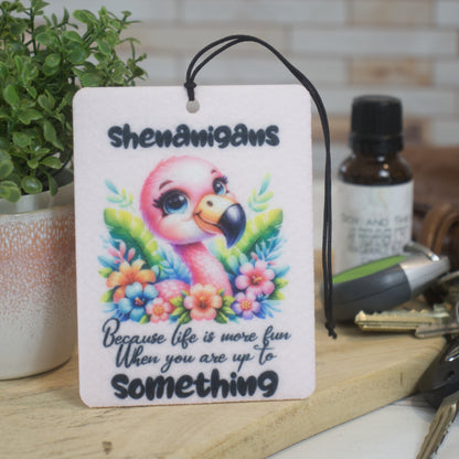 A car air freshener rests against a pot plant with the image of a cartoon flamingo surrounded by flowers and the words SHENANIGANS BECAUSE LIFE IS MORE FUN WHEN YOU ARE UP TO SOMETHING.  Behind is a bottle of scent and some car keys.