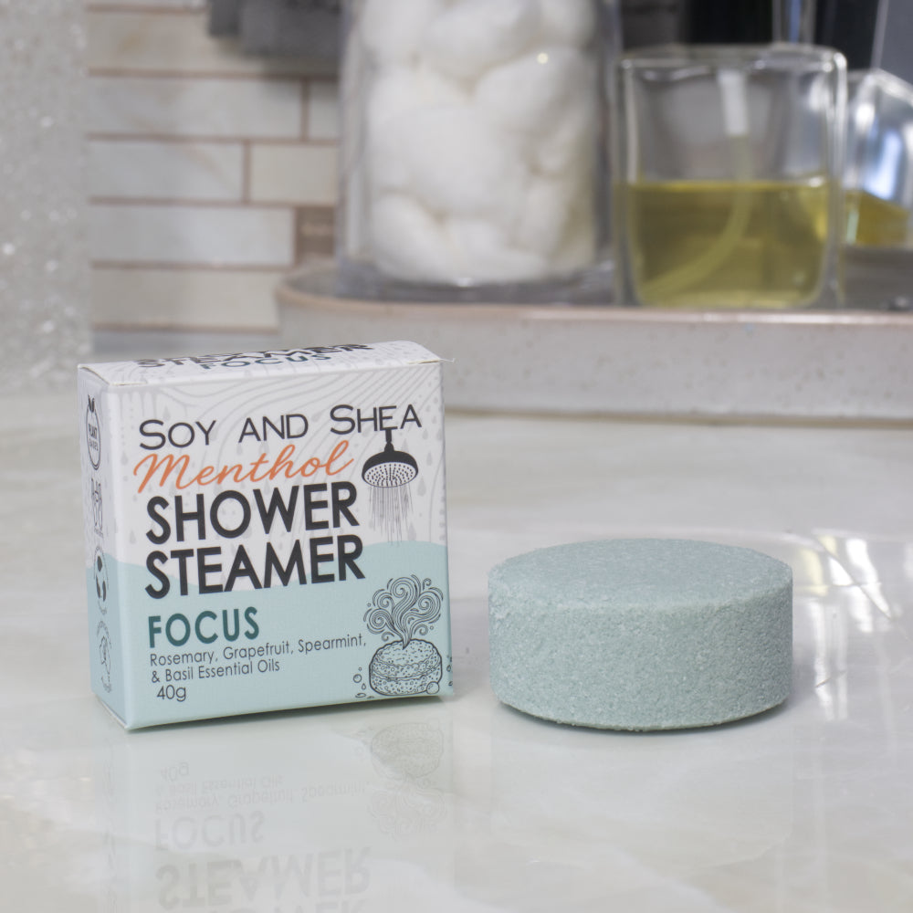 A boxed shower steamer and the steamer itself on a countertop in a bathroom setting. The box reads 'Soy and Shea Menthol Shower Steamer Focus' and the round steamer is a pale dark green color.