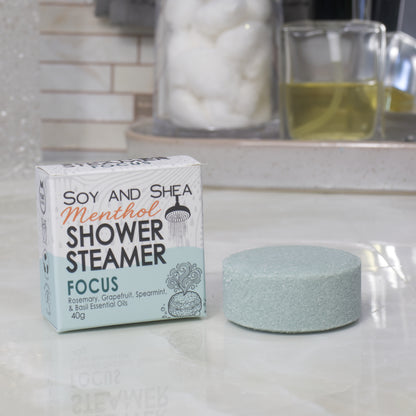 A boxed shower steamer and the steamer itself on a countertop in a bathroom setting. The box reads 'Soy and Shea Menthol Shower Steamer Focus' and the round steamer is a pale dark green color.