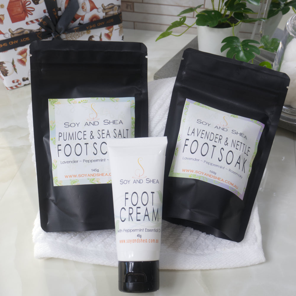 The Soy and Shea Mini Foot Pamper Set features black pouches of Pumice & Sea Salt and Lavender & Nettle foot soaks, plus a foot cream tube on a white towel—perfect for an at-home spa. Bathroom accessories finish this thoughtful gift.