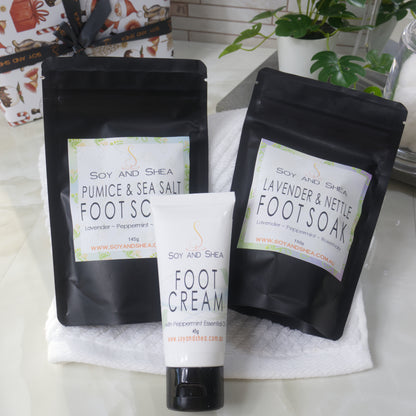 The Soy and Shea Mini Foot Pamper Set features black pouches of Pumice & Sea Salt and Lavender & Nettle foot soaks, plus a foot cream tube on a white towel—perfect for an at-home spa. Bathroom accessories finish this thoughtful gift.