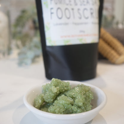 A small white bowl of green colours salts silts on a white surface with a blurred backfround including the pouch of Foot Scrub