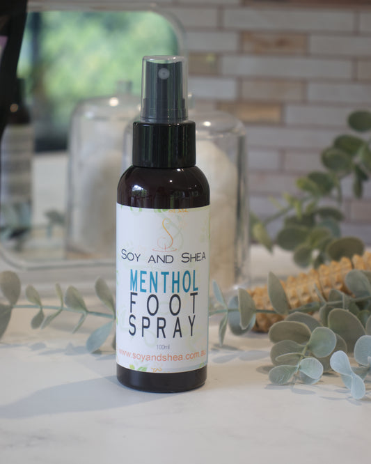 An amber bottle with atomiser of 'Soy and Shea Menthol Foot Spray' on a marble surface with decorative plants in the background.