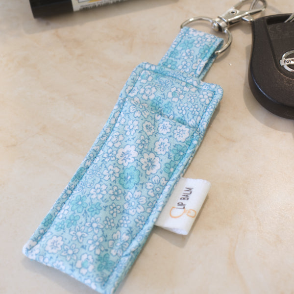 A clip balm holder lies on a wooden surface attached to a car key with a "lobster claw" clip. The Clip Balm is light blue and is covered in white and teal flowers.